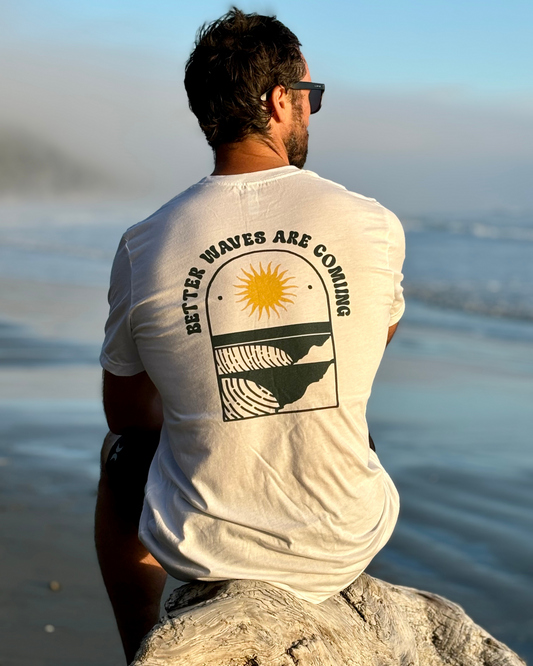 A man wearing sunglasses and a graphic surf shirt with the text "Better Waves Are Coming," sits on a log on a sandy beach, facing the ocean.