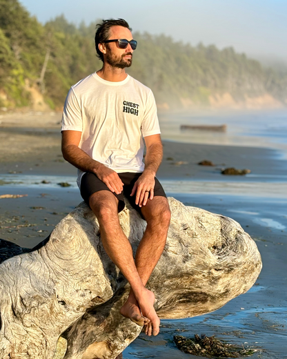 A man in a chest high surf graphic t-shirt and black shorts sits on a large driftwood log on a beach, with trees and mist in the background. He wears sunglasses and looks into the distance.